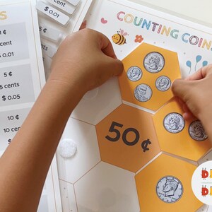 May include: A child's hands are placing US coins on a honeycomb-shaped board with the text "COUNTING COINS" and "50&cent;" printed on it. The board is white with yellow hexagons.