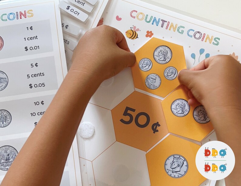 May include: A child's hands are placing US coins on a honeycomb-shaped board with the text "COUNTING COINS" and "50&cent;" printed on it. The board is white with yellow hexagons.