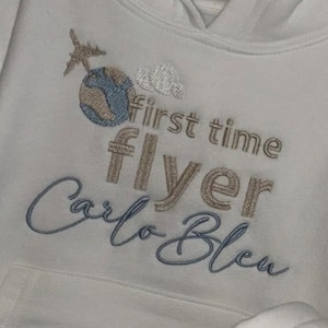 Könnte beinhalten: Ein weißer Kapuzenpullover mit gestickten Motiven in beigem und blauem Garn. Die Stickerei zeigt ein kleines Flugzeug, einen Globus und Wolken über dem Text "first time flyer" und dem Namen "Carlo Bleu" in einer geschwungenen Schreibschrift.