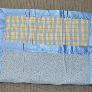 May include: A blue baby blanket with a plaid pattern on one side and a blue and white pattern on the other side. The blanket has a blue satin border.