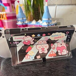 以下が含まれることがあります： 白のニンテンドースイッチ本体に、透明な保護ケースが装着されています。ケースには、黒と白のストライプ柄が施され、漫画のウサギと「Funny」と「Rabbit Rabbit」の文字が描かれています。