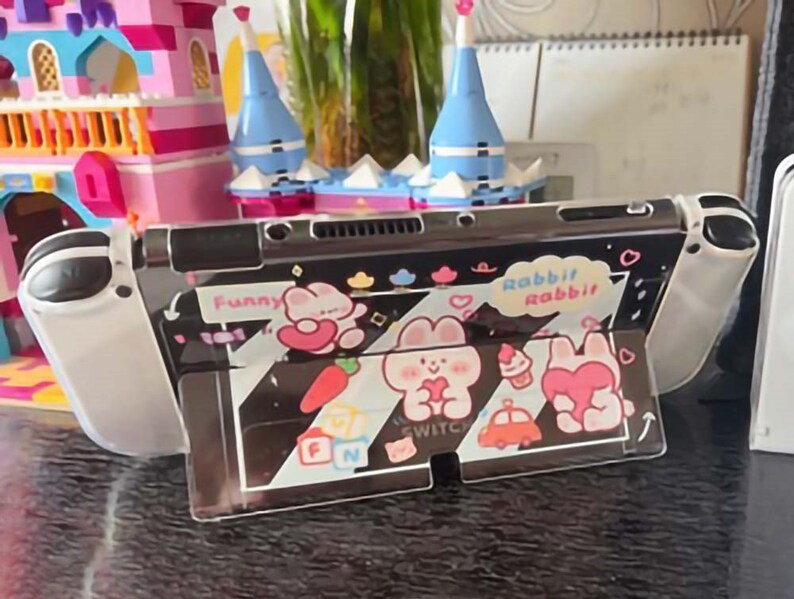 以下が含まれることがあります： 白のニンテンドースイッチ本体に、透明な保護ケースが装着されています。ケースには、黒と白のストライプ柄が施され、漫画のウサギと「Funny」と「Rabbit Rabbit」の文字が描かれています。