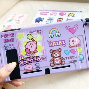 May include: A purple Game Boy Advance SP console with a variety of colorful stickers, including a rainbow, hearts, and a pink character. The console has a screen that displays a pink character with a star and the text "GAME BOY" and "カービィの きらきら".