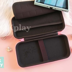 Puede incluir: Estuche de transporte rosa para una consola Nintendo Switch. El estuche est&aacute; abierto y tiene un interior negro con un bolsillo de malla. El estuche tiene las palabras "Ok Play" impresas en la parte delantera.