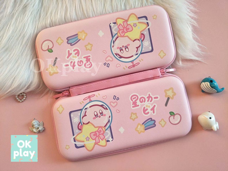 Puede incluir: Estuche de transporte rosa para Nintendo Switch con un dise&ntilde;o de personaje de dibujos animados que presenta un personaje rosa con una estrella y un melocot&oacute;n. El estuche tiene texto japon&eacute;s que dice "星のカーヒイ" que se traduce como "Kirby's Star".