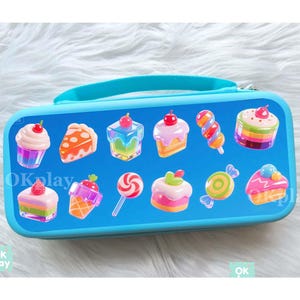 May include: A blue rectangular pencil case with a handle, decorated with cartoon sweets. The case features images of cupcakes, cake slices, lollipops, and candies. The text "OK play" is visible on the case.