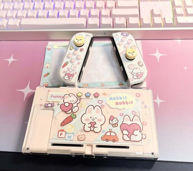 以下が含まれることがあります： ピンクと白のニンテンドースイッチ本体に、カスタムケースが装着されています。ケースはピンクの背景に白い水玉模様で、ハートを持った漫画のウサギが描かれています。ケースには白い雲の中に「Rabbit Rabbit」という文字が書かれています。本体には、カップケーキ、車、ニンジンなど、他の漫画の画像も飾られています。