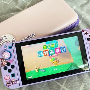 May include: A purple Nintendo Switch console with a pink and white case. The console has a screen displaying a video game with Japanese text. The console has custom buttons with paw prints and cartoon characters.