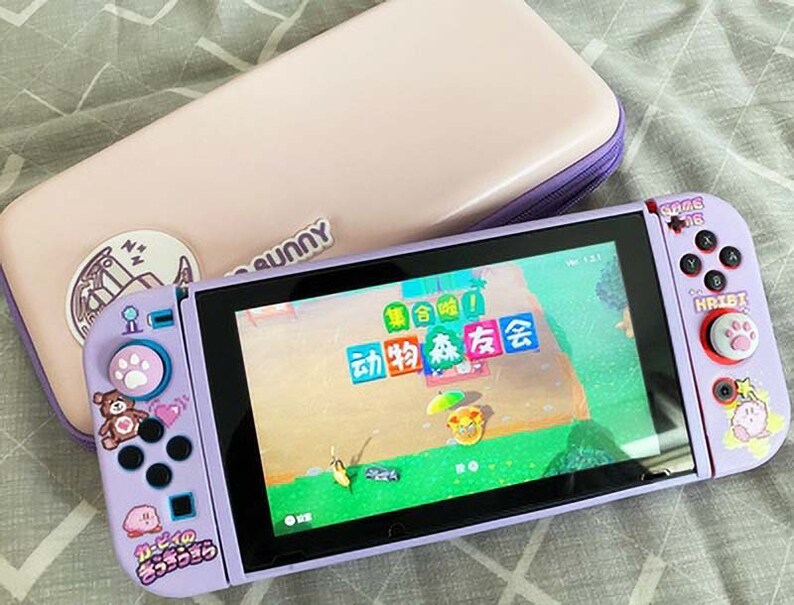 May include: A purple Nintendo Switch console with a pink and white case. The console has a screen displaying a video game with Japanese text. The console has custom buttons with paw prints and cartoon characters.