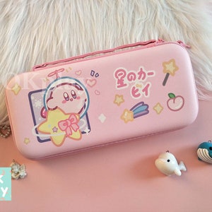 Puede incluir: Estuche rosa para Nintendo Switch con un personaje de dibujos animados de Kirby sosteniendo una estrella. El estuche tiene una frase japonesa que dice "星のカービィ" y otros dise&ntilde;os de estrellas y corazones.