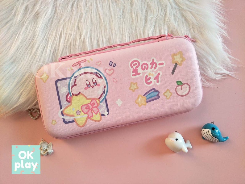 Puede incluir: Estuche rosa para Nintendo Switch con un personaje de dibujos animados de Kirby sosteniendo una estrella. El estuche tiene una frase japonesa que dice "星のカービィ" y otros dise&ntilde;os de estrellas y corazones.
