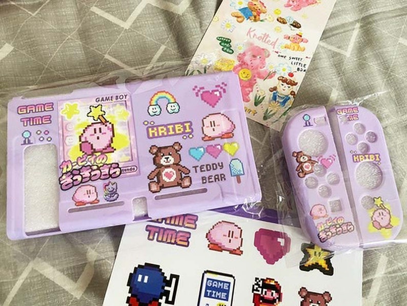 May include: A purple and pink Nintendo Switch game case with a pink Kirby character design. The case has a matching controller skin with a pink Kirby character design. The case and controller skin have the text "Game Time" and "Kribbi" on them.