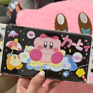 Può includere: Una console Nintendo Switch bianca con una custodia trasparente che presenta un personaggio rosa e bianco di Kirby, un personaggio giallo di Waddle Dee e il testo giapponese "カービィ". La custodia ha uno sfondo nero con stelle, cuori e altri elementi decorativi.