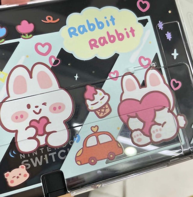 以下が含まれることがあります： 透明なプラスチックカバーが付いた黒いニンテンドースイッチケース。ケースには、ピンクのハートを持った白いウサギ2匹と、背中にピンクのハートをつけた白いウサギが描かれた、漫画のデザインが施されています。ケースには、赤い車、ピンクのアイスクリームコーン、黄色の吹き出しの中に「Rabbit Rabbit」という文字も描かれています。