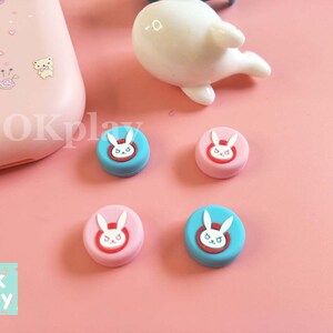 以下が含まれることがあります： 白いウサギのデザインが施された、丸いカラフルなシリコン製ゲームコントローラーのサムスティックキャップ4個。キャップはピンクとブルーです。