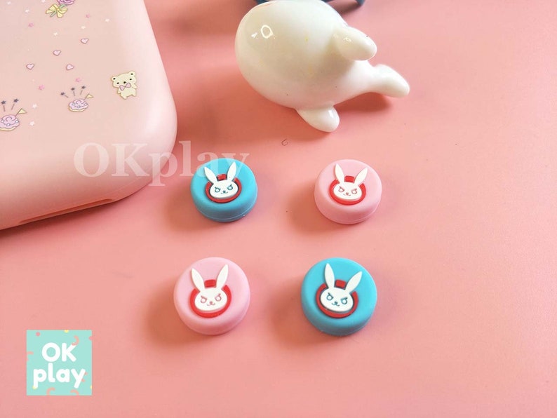 以下が含まれることがあります： 白いウサギのデザインが施された、丸いカラフルなシリコン製ゲームコントローラーのサムスティックキャップ4個。キャップはピンクとブルーです。