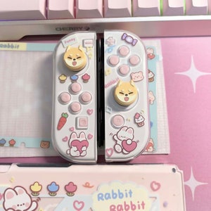 以下が含まれることがあります： ピンクと白のデザインの白いニンテンドースイッチジョイコンコントローラー。コントローラーには、ジョイスティックに漫画の犬の顔、ボタンに漫画のウサギの顔が描かれています。コントローラーは、ハート、ニンジン、カップケーキで飾られています。