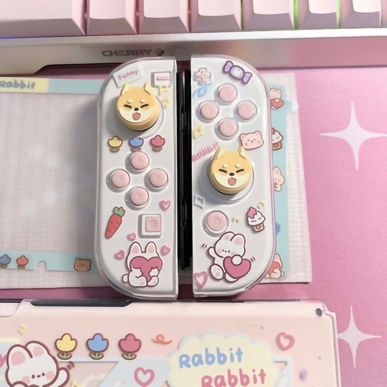 以下が含まれることがあります： ピンクと白のデザインの白いニンテンドースイッチジョイコンコントローラー。コントローラーには、ジョイスティックに漫画の犬の顔、ボタンに漫画のウサギの顔が描かれています。コントローラーは、ハート、ニンジン、カップケーキで飾られています。