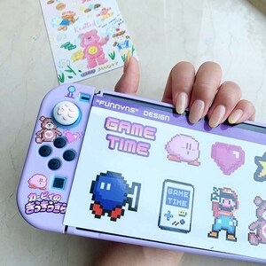May include: A purple Nintendo Switch console with a custom design featuring pixelated characters and the text "FUNNYNS" DESIGN GAME TIME". The console has a pink button with a heart design and a pink and white Kirby character sticker.