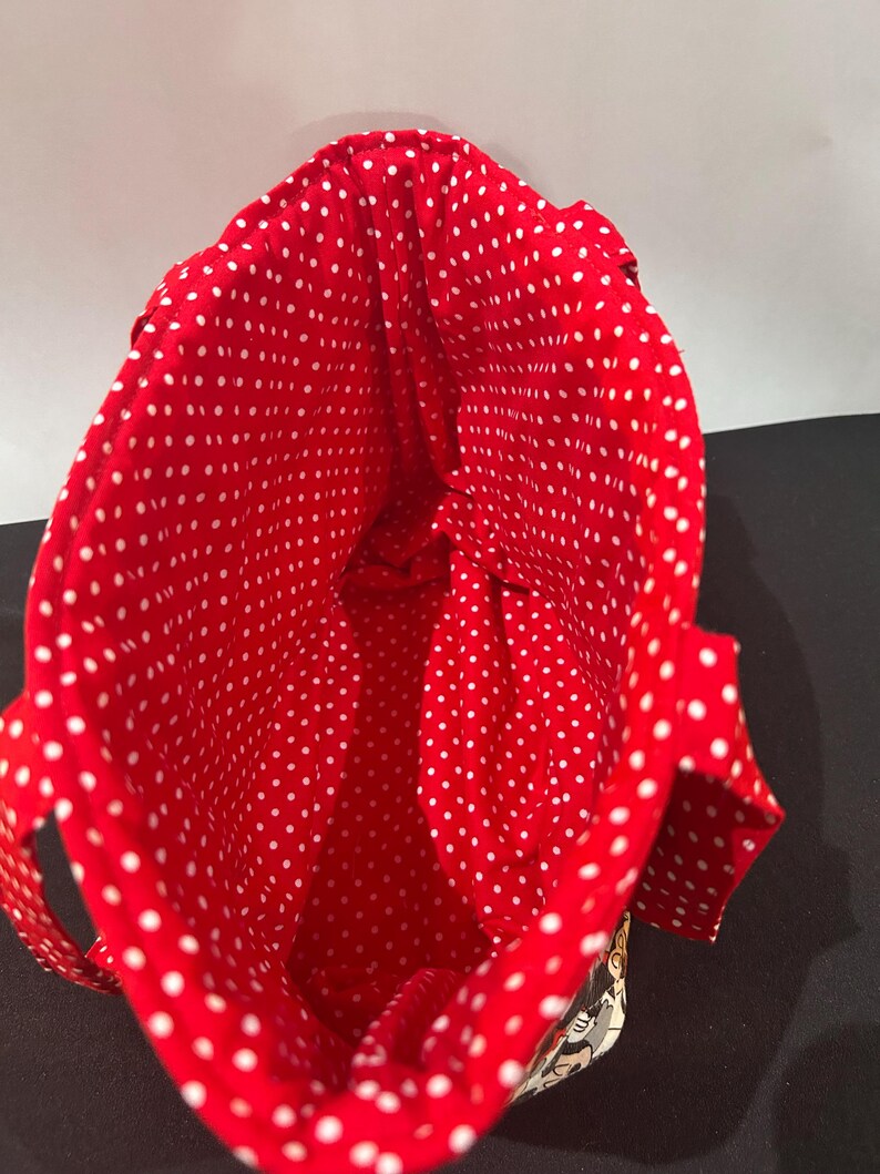 May include: A fabric tote bag featuring a vibrant red interior lining with a white polka dot pattern. The bag has two matching red and white polka dot fabric handles attached to the sides, with a glimpse of a patterned exterior visible at the base.