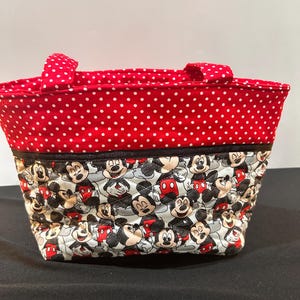May include: A quilted fabric tote bag featuring a top section of red fabric with white polka dots and a bottom section with a repeating pattern of Mickey Mouse characters. The bag includes two matching red and white polka dot fabric handles.