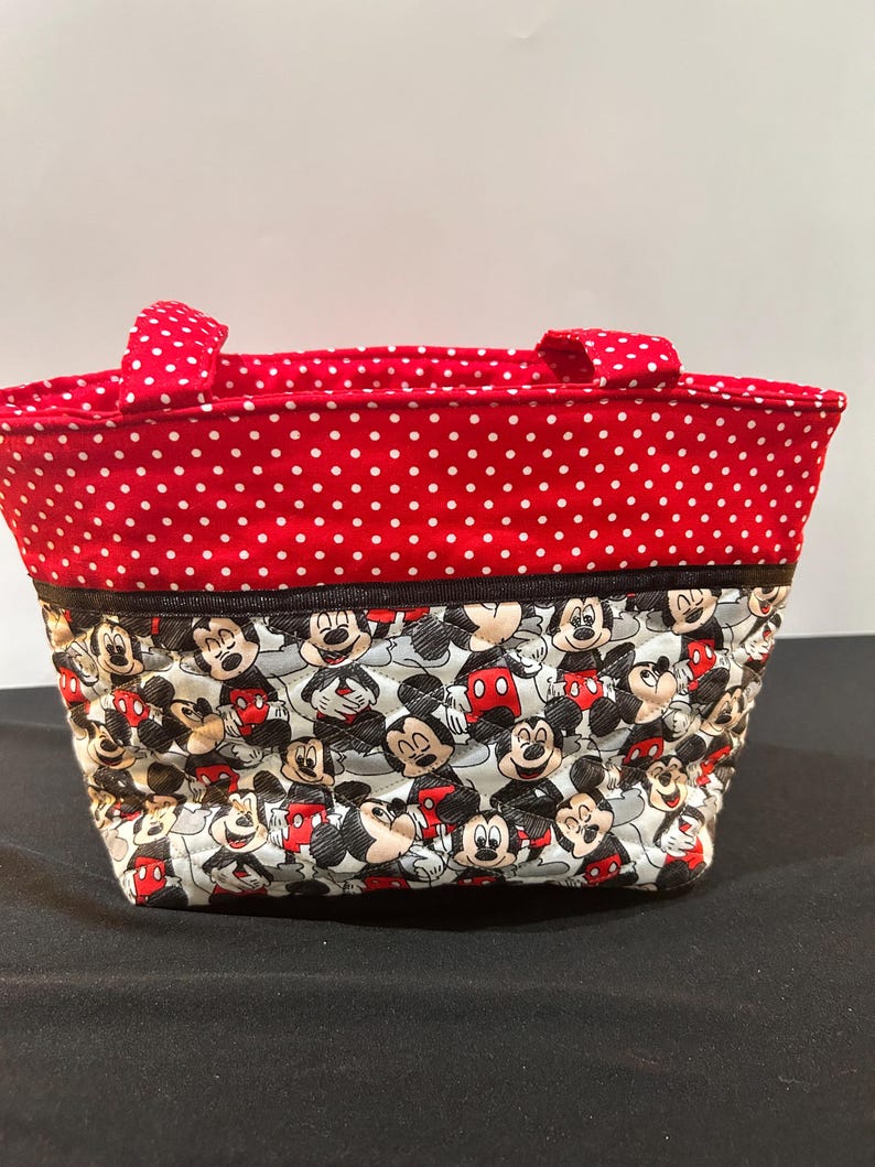 May include: A quilted fabric tote bag featuring a top section of red fabric with white polka dots and a bottom section with a repeating pattern of Mickey Mouse characters. The bag includes two matching red and white polka dot fabric handles.