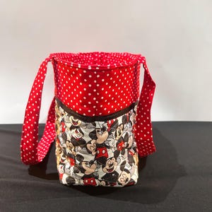 May include: A quilted fabric bucket bag featuring a repeating Mickey Mouse character print on the lower section and a red fabric with white polka dots on the upper section and handles. The bag has a dark brown trim separating the two fabric patterns.