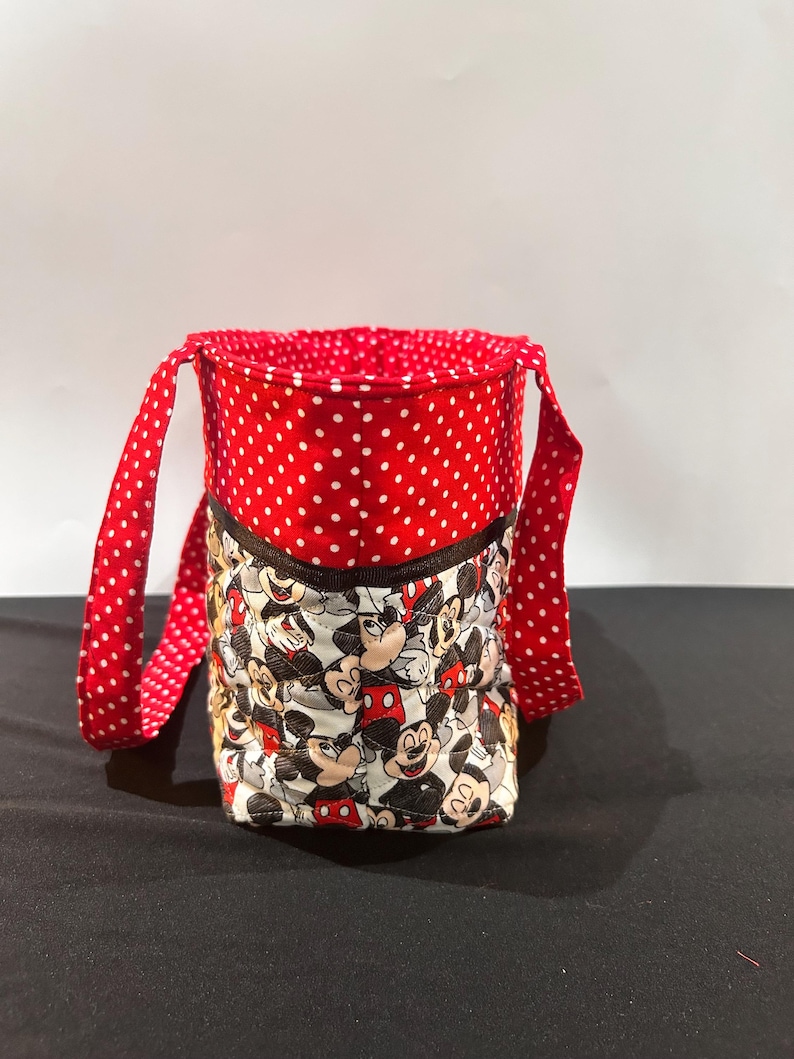 May include: A quilted fabric bucket bag featuring a repeating Mickey Mouse character print on the lower section and a red fabric with white polka dots on the upper section and handles. The bag has a dark brown trim separating the two fabric patterns.
