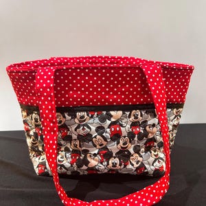 May include: A quilted tote bag featuring a repeating pattern of Mickey Mouse characters on a white background. The upper portion of the bag and the two shoulder straps are made of red fabric with a white polka dot pattern. A black trim separates the sections.