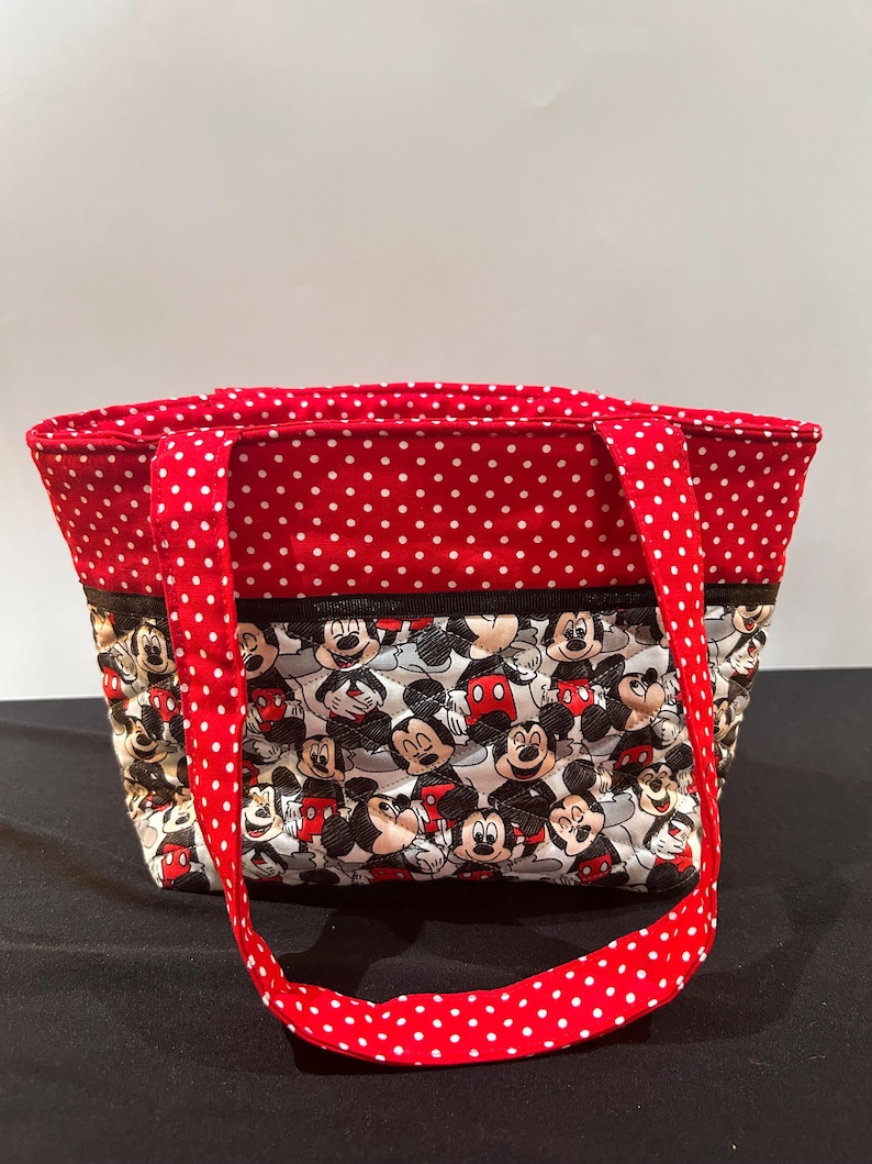 May include: A quilted tote bag featuring a repeating pattern of Mickey Mouse characters on a white background. The upper portion of the bag and the two shoulder straps are made of red fabric with a white polka dot pattern. A black trim separates the sections.