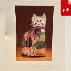 Pode incluir: Um brinquedo de gato de patchwork feito de vários tecidos em tons de verde, vermelho e marrom. O gato tem um rosto branco com olhos pretos e uma boca vermelha.