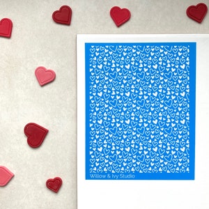 May include: A blue stencil with a white pattern of many hearts. The stencil is on a white surface with red hearts scattered around it. The text "Willow & Ivy Studio" is at the bottom of the stencil.
