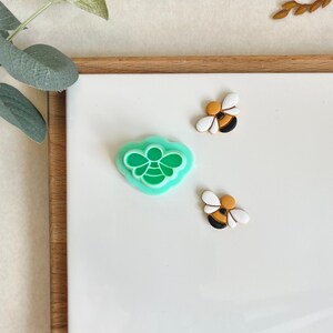 May include: A green plastic bee cookie cutter and two small bee-shaped decorations with white and brown accents.