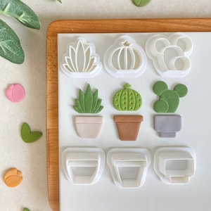 May include: A set of 10 white plastic cookie cutters shaped like succulents and pots. The cutters are on a white surface with green, brown, and grey fondant succulents in the pots.