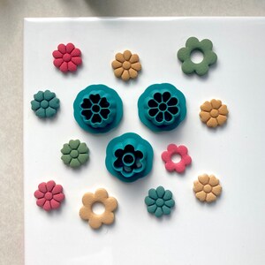 May include: A set of teal flower-shaped cookie cutters with a variety of colourful flower-shaped cookies in pink, green, and yellow.