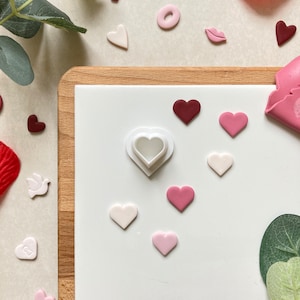 May include: A white heart-shaped cookie cutter on a white surface with pink and red heart-shaped fondant pieces.