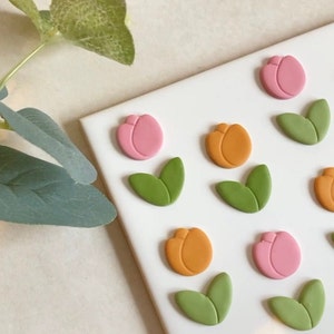 May include: A white surface with six pink and orange tulip-shaped clay decorations and six green leaf-shaped clay decorations. There are two white plastic cookie cutters in the bottom left corner of the image.