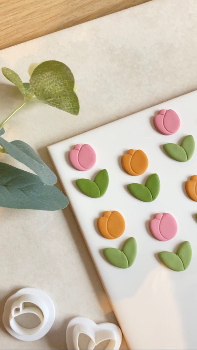 May include: A white surface with six pink and orange tulip-shaped clay decorations and six green leaf-shaped clay decorations. There are two white plastic cookie cutters in the bottom left corner of the image.