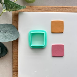 May include: A turquoise square-shaped cookie cutter with a smaller square cutout inside. There are two squares of polymer clay, one orange and one pink, on a white surface.