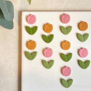 May include: A white surface with 12 handmade clay flower embellishments. The flowers are in three colours: pink, orange, and green. Each flower has a single bloom and two leaves.