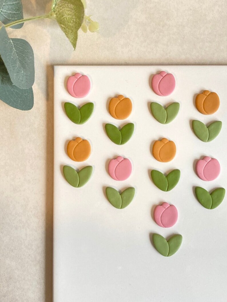 May include: A white surface with 12 handmade clay flower embellishments. The flowers are in three colours: pink, orange, and green. Each flower has a single bloom and two leaves.