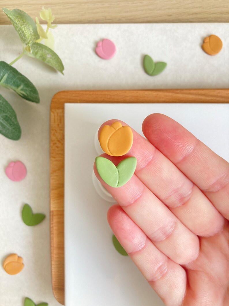 May include: A set of two polymer clay flower charms. One charm is a yellow tulip and the other is a green leaf.