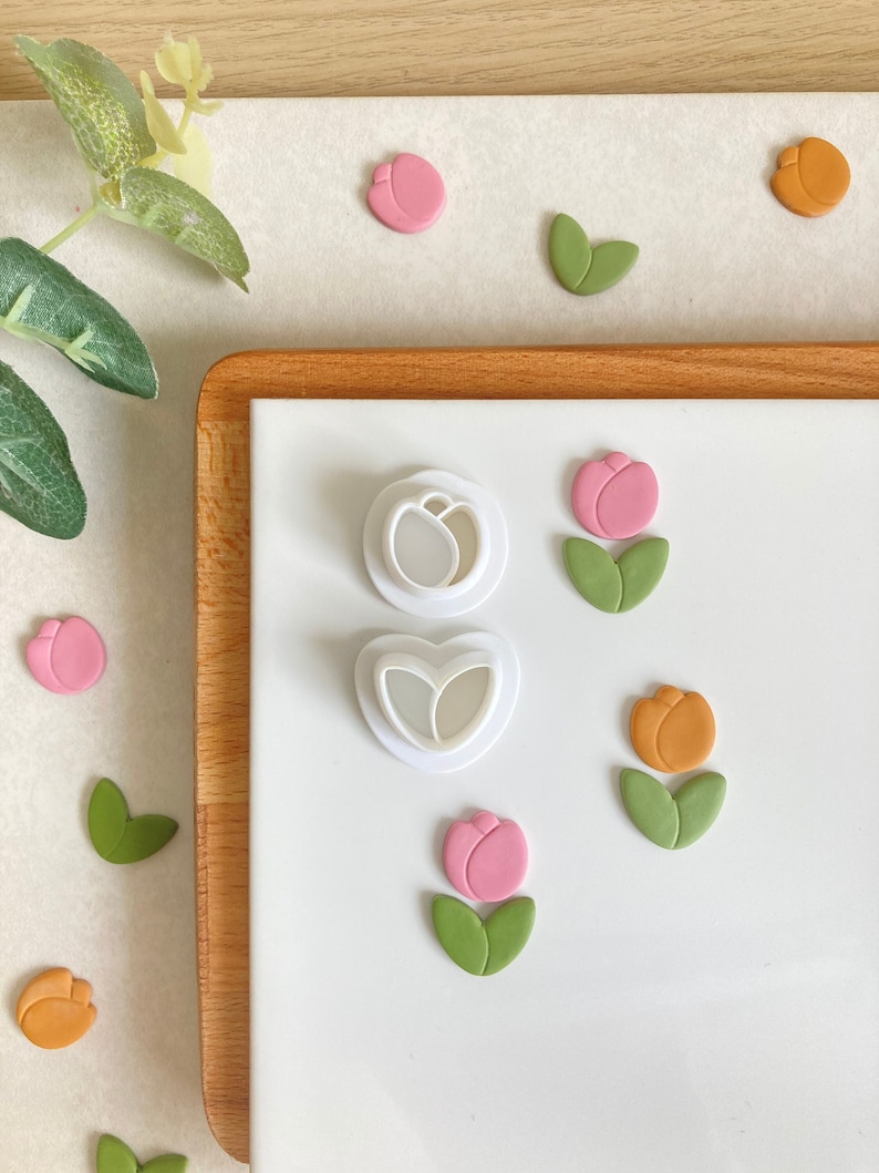 May include: Two white plastic cookie cutters shaped like tulips, one with a single tulip and the other with two tulips. There are also several pink and orange fondant tulips on a white surface.