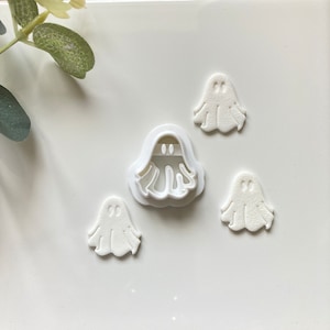 May include: Four white ghost-shaped cookie cutters, including one larger cutter with a handle. The cutters are on a white surface.