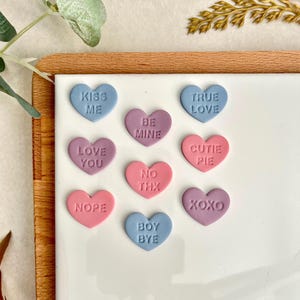 May include: A selection of pastel-coloured heart-shaped sweets, each bearing a different romantic message such as "Kiss Me" and "Be Mine". The sweets are displayed on a white surface, ideal for Valentine's Day or other celebrations of love.