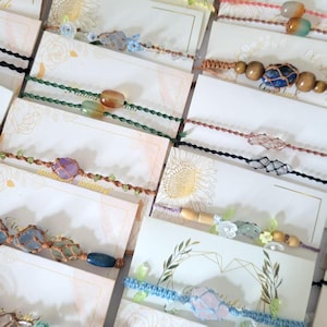 May include: Selection of handmade bracelets on display cards. The bracelets incorporate gemstones, beads, and macrame knots in colours such as blue, green, purple, and brown. Each bracelet is unique, showcasing different stone shapes and sizes.