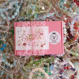 Puede incluir: Una caja de regalo rosa atada con hilo rosa y una pegatina con dibujos de gatos. La caja está rodeada de una variedad de pulseras y collares de cuentas de colores, creando una exhibición vibrante.