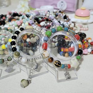 May include: A collection of colorful beaded bracelets displayed on clear stands. The bracelets feature a variety of beads in different colors, shapes, and sizes, including round, star-shaped, and patterned beads. Some bracelets have dangling charms.