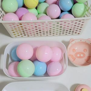 May include: A selection of pastel-coloured plastic balls in white baskets and containers. The balls are pink, blue, green, and yellow. A small pink pig-shaped bowl is also visible.