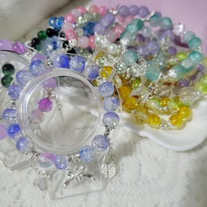 May include: A collection of colorful beaded bracelets in various shades of pink, purple, blue, green, and yellow. Some bracelets have silver charms, including bows and floral designs. The bracelets are displayed on a white surface.